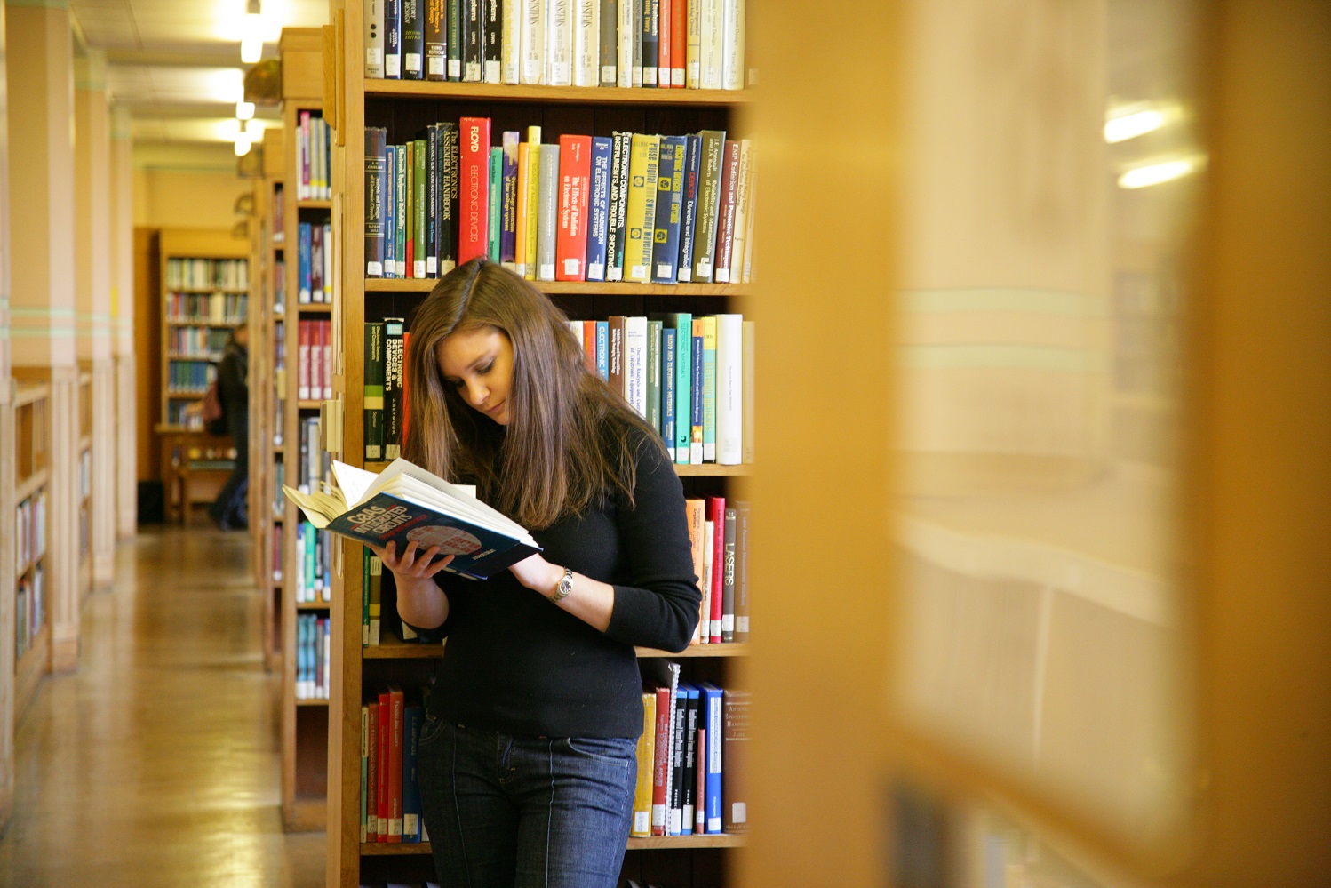 Shows a girl reading a book, in the background are stacks of shelves filled with books.