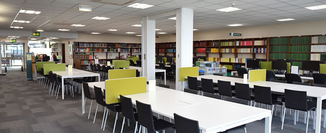 Large open plan room of the Chemistry Library, around the walls are bookshelves filled with books and in the middle of the room are rows of large desk tables with chairs.