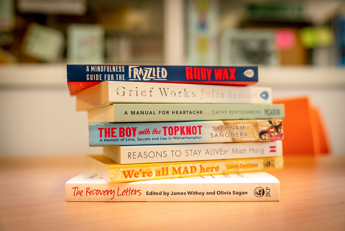 Stack of wellbeing books with a blurred background. Titles include, for example, 'Reasons to stay alive- Matt Haig' and 'A mindfulness guide for the frazzled - Ruby Wax'
