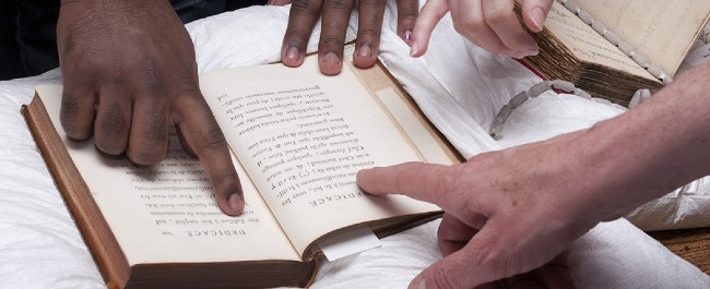 An open book on a cushion with hands pointing at the text.