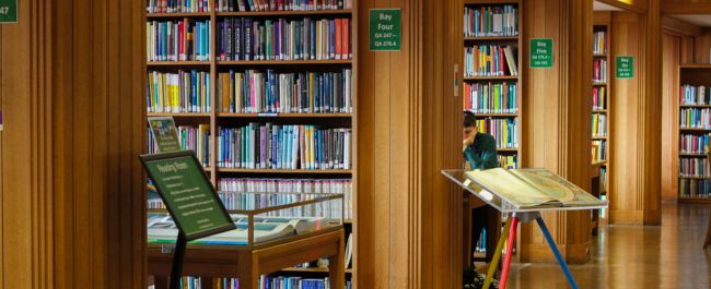 Inside of the Queens library. There are old wooden shelves full of multicoloured books, and a student studying at a desk. There is also a large David Hockney art book and display cabinet between the shelves.