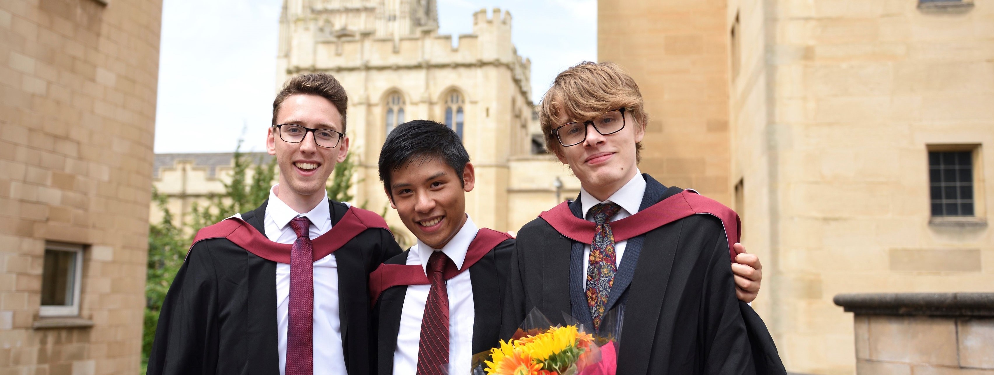 Three students in graduation gowns smile for the camera. A section of the Wills Memorial building is visible in the background. One of the graduands is holding a bouquet of flowers.