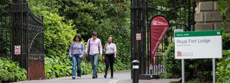 Three people walking through the gates of Royal Fort Gardens. They appear to be in conversation. In the foreground there is sign which says 