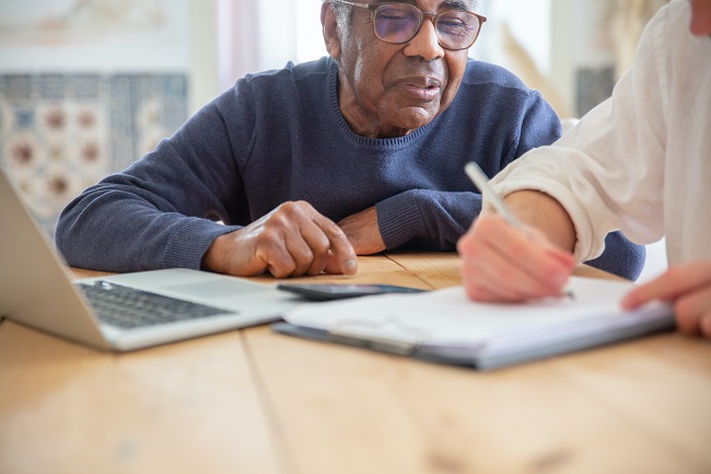 Older Black male care home resident sits at table with White male carer who is taking notes.