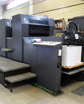 Image showing a grey and black Litho Printing press loaded up at one end with a large stack of paper ready to print, a stepped walkway is along the side for machine access