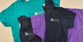 Image showing multiple University of Bristol branded t shirts and Polos with the logo printed on the front left breast, T shirt colours are ranging from Aqua to the left hand side to Navy Blue and Purple on the right of the image