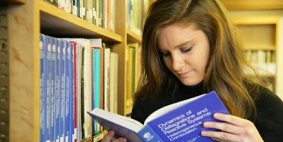 A woman reading from a book in a library. 
