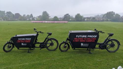 Two University of Bristol Sport Exercise and Health electric black cargo bikes positioned on grass next to cricket pitches. The cargo bikes have a large space at the front for you to place items in for transport and are branded with the text 'Future proof, helping to deliver a net zero campus' on their side. 