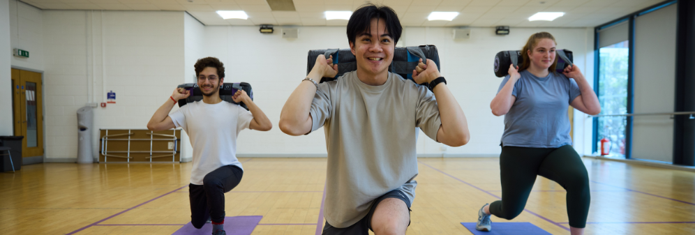 Three students stretching in a fitness studio with weighted bags on their upper backs