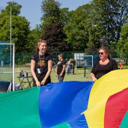 Lucy laughing holding part of a colourful parachute with other people on a sport fields with additional sport activities taking place in the background.