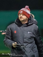 Image of a man wearing a padded coat and woolly hat with an ipad in his hands. He is coaching hockey on a pitch