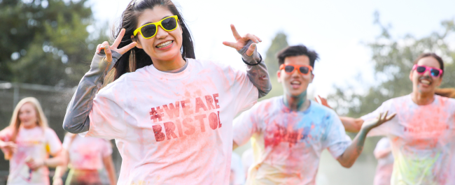 Female and male students smiling and posing for the camera, wearing a white #WeAreBristol t-shirt that is covered in different coloured powder paint. Each person is wearing colourful sunglasses