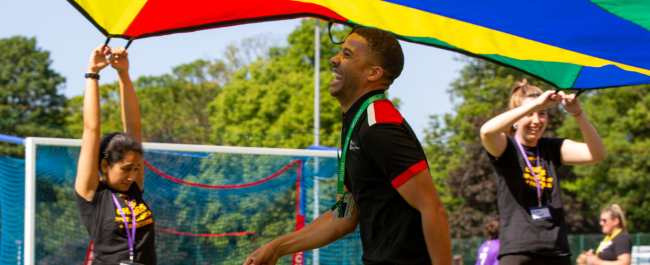 Two Game Changers holding up a colourful parachute as a member of staff walks under neath it laughing and smiling.