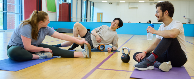 Three smiling students sitting on a gym studio floor in front of a mirrored wall each performing a different action. A female is stretching their left leg, one male is using a foam roller on their lower back and another male is sitting crossed legged with a water bottle.