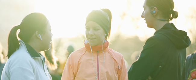 Three females standing facing one another wearing headphones and athletic clothing.