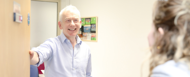 Osteopath Gerry smiling holding open a treatment room door as a client walks towards him.