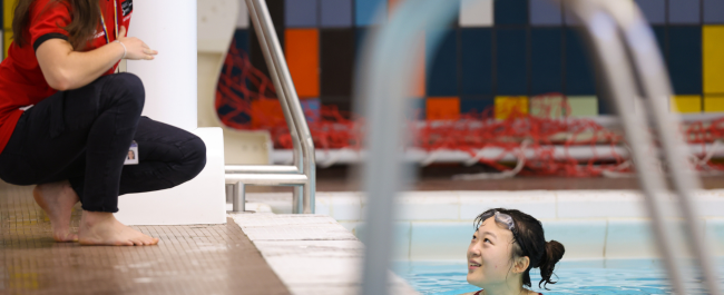 A lifeguard who is poolside speaking to a swimmer in the water.