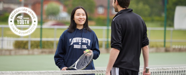 LTA Youth Recognised Venue stamp overlaying an image of a female holding a tennis racket and ball speaking to male at the court net.