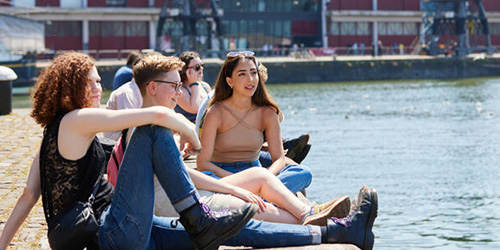 3 students sitting by Bristol harbour on a sunny day