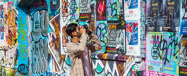A male student taking a photo of a wall covered in posters and graffiti