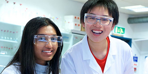 two young students in lab coats and protective goggles smiling at the camera