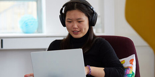 A happy female student sat on a chair with headphones looking at a laptop