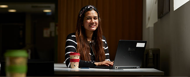 A student sits with their laptop and a hot drink.