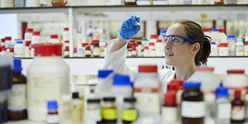 A student dressed in lab coat and goggles examines a test tube