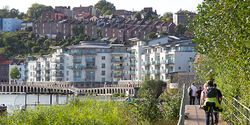 People walking and cycling over a bridge at the harbourside. A large number of terraced houses and apartments sit on the hills behind.