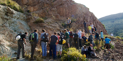A group of people with backpacks and hard-hats consulting a large rock face.
