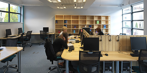 Three students working independently in a University study space surrounded by computers, sets of headphones, and books.