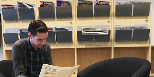 A student reading a newspaper in front of shelves containing newspapers written in various languages.