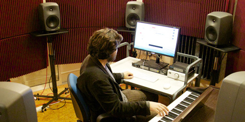 A person working in a sound recording studio with a keyboard, computer and large speakers surrounding them.