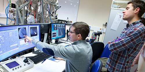 Two people working at a computer in a laboratory with a large machine in the background.