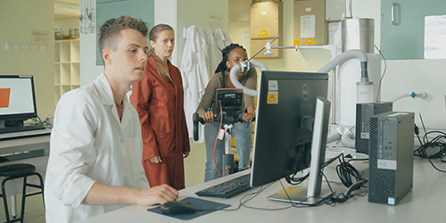 Three people in a laboratory. One is in a white lab coat working at a computer, while one is riding an exercise bike and another stands nearby looking at the computer.