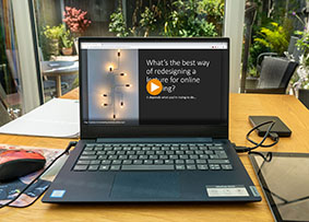 A mediasite video about redesigning a lecture for online teaching on a laptop. On the table the laptop is on, there is also a tablet, a mouse, a hard drive and sheets of paper. A garden is visible in the background.