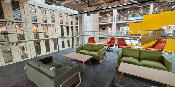 Modern green sofas and orange chairs arranged around low coffee tables, surrounded by a glass balcony. The lounge overlooks a view of the inside of an industrial building.