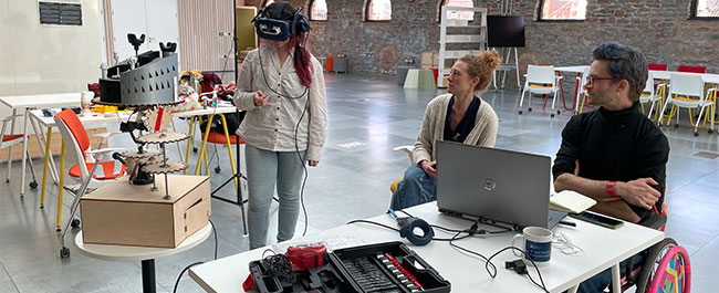 A person standing with a Virtual Reality headset on and two people sitting at a table with a laptop.