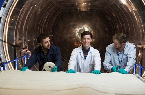 Three people in green gloves with tape measure and scissors standing, smiling, at a trolley with huge sheets of material on, in the mouth of a huge silver tunnel.