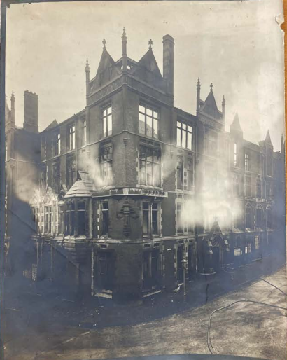 Black and white photograph of the corner of a large four storey building, with smoke coming out of several windows.
