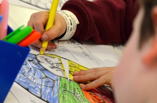 Children in burgundy uniform sit at a table. A child's hand grips a colouring pencil and is colouring in a worksheet.