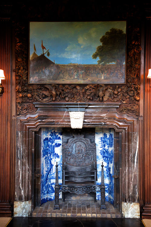 A large fire place with painted tile surround and Intricately carved wood decoration