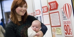 A young woman looks at Children of the 90s poster display holding a baby.